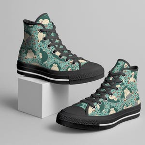 Op de afbeelding: Hoge sneakers met een teal achtergrond en een herhaald patroon van een stripfiguur. De schoenen hebben zwarte zolen, veters en een witte streep. De schoenen worden gepresenteerd op een witte kubus.