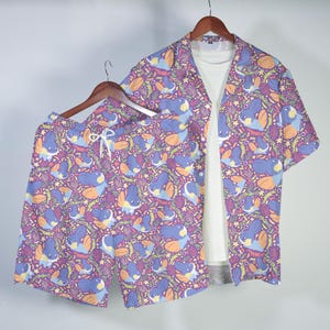 May include: A matching purple short-sleeve shirt and shorts set. The set features a repeating pattern of blue, orange, and yellow sea creature illustrations on a purple background. The shorts have a white drawstring. The shirt is open, revealing a white t-shirt.
