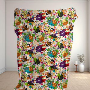 Op de afbeelding: Een rechthoekige deken met een herhalend patroon van diverse cartoonwezens, oranje pompoenen, groene bladeren en kleine kleurrijke bloemen op een witte achtergrond. Het ontwerp bevat paarse, blauwe, gele en bruine figuren.