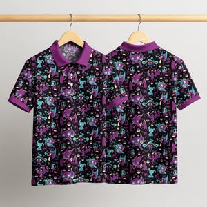 May include: Two short-sleeved polo shirts with a black background and a repeating pattern of purple, teal, and white cartoon characters. The shirts have purple collars and sleeve cuffs, and are hanging on a wooden hanger.