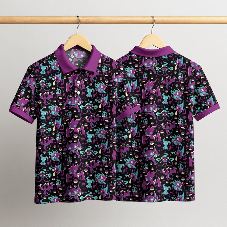 May include: Two short-sleeved polo shirts with a black background and a repeating pattern of purple, teal, and white cartoon characters. The shirts have purple collars and sleeve cuffs, and are hanging on a wooden hanger.