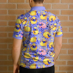 May include: A short-sleeved polo shirt with a repeating pattern of cartoon sheep in yellow, blue, and pink on a lavender background. The shirt has a light blue collar and sleeve cuffs. The person is standing in front of a brick wall.