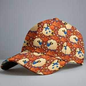 May include: A baseball cap featuring a repeating pattern of cartoon sheep, moons, and stars on a rust-coloured background. The cap has a curved brim and a button on top. The sheep have blue faces and yellow accents.
