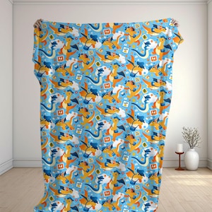 Puede incluir: Una manta de color azul claro con un patrón repetitivo de criaturas naranjas parecidas a dragones que llevan sombreros y bolsos azules. El diseño incluye pequeñas nubes blancas, iconos cuadrados naranjas con palmeras y corazones rojos.