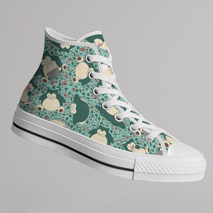 Op de afbeelding: Hoge canvas sneaker met een turquoise achtergrond met een herhaald patroon van stripfiguren, harten en andere kleine vormen. De sneaker heeft witte veters, een witte rubberen zool en een zwarte streep.
