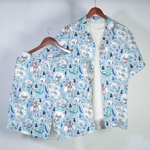 May include: A short-sleeved button-down shirt and matching shorts set in light blue, featuring an all-over print of various cartoon creatures, pine trees, and snowflakes. The shirt is layered over a plain white t-shirt on a wooden hanger.