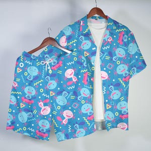May include: A matching set of blue shorts and a short-sleeved button-up shirt. Both items feature a repeating pattern of cartoon characters, skateboards, and geometric shapes in pink, yellow, and blue. The shirt is open, revealing a white t-shirt underneath.