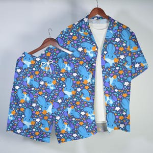 May include: A short-sleeve button-down shirt and matching shorts set featuring a dark blue background with a repeating pattern of light blue cartoon creatures, orange stars, and white star shapes, worn over a plain white t-shirt.