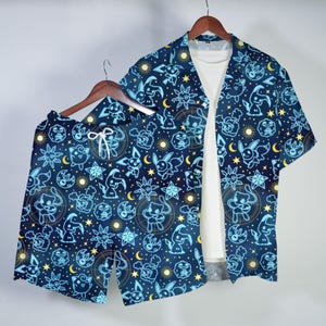 May include: A navy blue shirt and shorts set with a repeating pattern of cartoon characters, moons, and stars in light blue and yellow. The short-sleeved shirt is open, revealing a white undershirt. The shorts have a drawstring waist. The set is hanging on a wooden hanger.