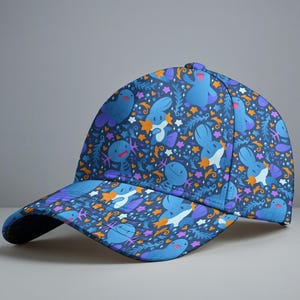 May include: A baseball cap with a navy blue background and a repeating pattern of cartoon-like blue creatures, orange stars, and purple accents. The cap has a curved brim and a button on top.