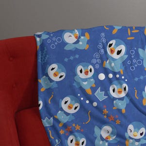 May include: A blue fleece blanket featuring a repeating pattern of cartoon penguins in various poses. The design includes small white snowflakes, orange starfish, yellow curved lines, and light blue crown shapes scattered across the fabric.