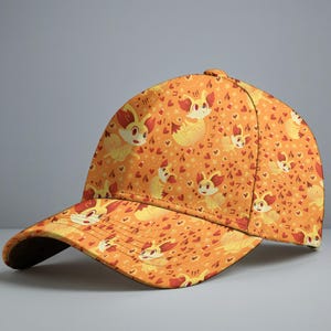 May include: Orange baseball cap featuring a repeating pattern of a cartoon fox-like creature, hearts, and Japanese text. The cap has a curved brim and a button on top. The design includes red, yellow, and white accents.