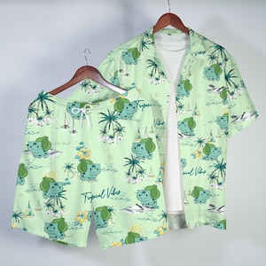 Könnte beinhalten: Hellgrünes Hemd- und Shorts-Set mit sich wiederholendem Cartoon-Charakter, Palmen und Segelboot-Muster. Das Hemd ist kurzärmlig und offen, darunter ein weißes T-Shirt. Die Shorts haben einen Kordelzug. Der Text "Tropical Vibes" ist aufgedruckt.