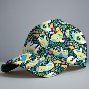 May include: A baseball cap with a whimsical pattern of yellow and blue cartoon creatures with closed eyes, white wings, and floral accents on a dark teal background. The cap has a curved brim and a structured crown.