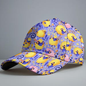 May include: A baseball cap with a playful design of yellow sheep on a lilac backdrop. The cap has a curved peak and a structured crown. The sheep are illustrated with blue details, alongside stars and floral motifs.