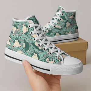 Op de afbeelding: Hoge canvas sneakers met een turquoise achtergrond en een herhaald patroon van een stripfiguur in verschillende poses. De sneakers hebben witte veters, een witte neus en een zwarte streep langs de zool.