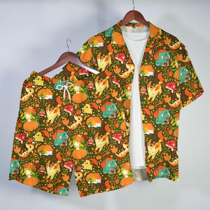Op de afbeelding: Een set bestaande uit een overhemd met korte mouwen en bijpassende korte broek, voorzien van een all-over print van diverse stripfiguren, pompoenen en herfstbladeren in oranje, groene en gele tinten, getoond over een effen wit T-shirt.