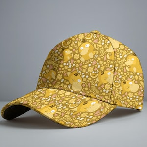 May include: A yellow baseball cap with a repeating pattern of cartoon characters, flowers, and fruit slices. The cap has a curved peak and a button on top. The design is in yellow, pink, and white on a khaki background.
