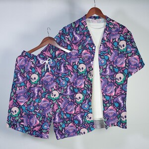 Puede incluir: Un conjunto a juego de camisa de manga corta y pantalones cortos morados. El conjunto presenta un patrón repetido de monstruos de dibujos animados, calaveras y sombreros de bruja en tonos de morado, rosa y verde azulado sobre un fondo azul oscuro. Los pantalones cortos tienen un cordón blanco.