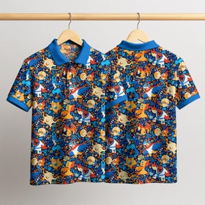 Puede incluir: Dos polos de manga corta con un estampado vibrante de personajes de dibujos animados en tonos azules, naranjas, amarillos y blancos. Las camisas tienen cuello y ribete de manga azules, colgando de una percha de madera.