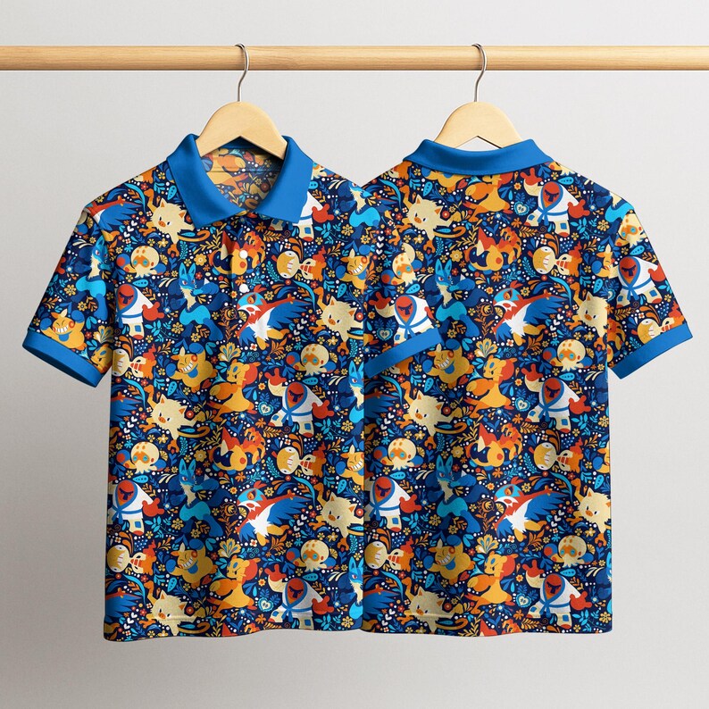 Puede incluir: Dos polos de manga corta con un estampado vibrante de personajes de dibujos animados en tonos azules, naranjas, amarillos y blancos. Las camisas tienen cuello y ribete de manga azules, colgando de una percha de madera.