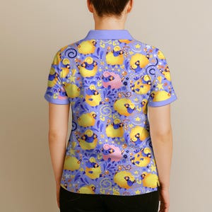 May include: A short-sleeved polo shirt with a whimsical pattern of yellow and pink sheep on a periwinkle background. The sheep are adorned with blue horns and are interspersed with stars and floral elements. The shirt has a light purple collar and sleeves.