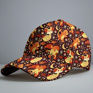 May include: A baseball cap with a repeating pattern of orange and yellow cartoon characters, moons, and stars on a dark brown background. The cap has a curved brim and a rounded crown.