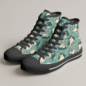 Op de afbeelding: Hoge canvas sneakers met een herhaald patroon van een stripfiguur in groen, beige en rood op een turquoise achtergrond. De sneakers hebben zwarte veters, zwarte zolen en een witte streep langs de zool.