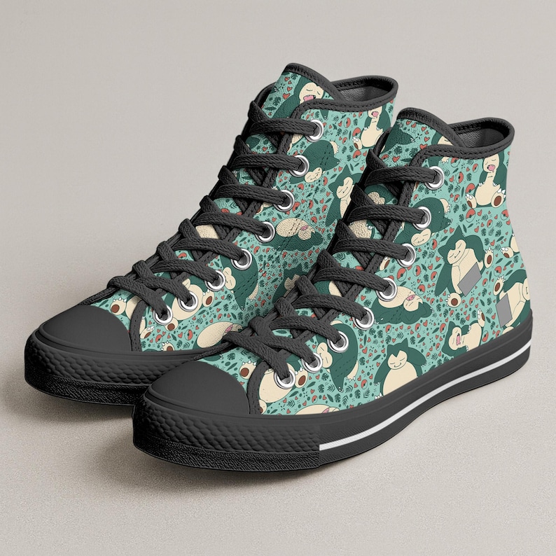 Op de afbeelding: Hoge canvas sneakers met een herhaald patroon van een stripfiguur in groen, beige en rood op een turquoise achtergrond. De sneakers hebben zwarte veters, zwarte zolen en een witte streep langs de zool.