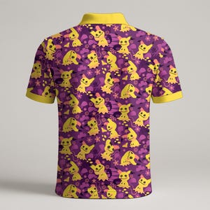 May include: A purple polo shirt with a yellow collar and sleeve trim. The shirt has a repeating pattern of yellow cartoon characters and floral elements. The design is set against a purple background.