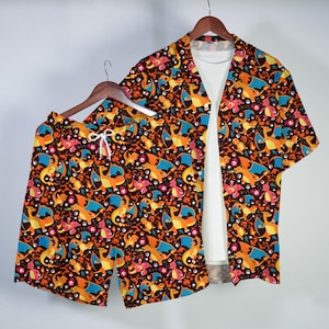 Op de afbeelding: Een bijpassende kledingset, bestaande uit een shirt met korte mouwen en een korte broek, beide met een herhalend patroon van oranje, blauwe en roze cartoonfiguren op een donkerbruine achtergrond. Het shirt is open en onthult een wit ondershirt.