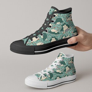 Op de afbeelding: Hoge canvas sneakers met een turquoise achtergrond en een herhaald patroon van een stripfiguur. De sneakers hebben zwarte zolen, veters en afwerking. De onderste sneaker heeft witte veters en afwerking. De stripfiguur is wit en groen.