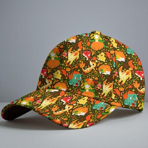 Op de afbeelding: Een baseballpet met een all-over print op een donkergroene achtergrond. Het patroon bevat diverse kleine cartoonfiguren, oranje pompoenen, rode paddenstoelen en herfstbladeren in tinten geel, oranje en groen over de gehele stof verspreid.