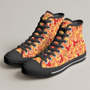 Könnte beinhalten: Hohe Sneaker mit einem sich wiederholenden Muster aus gelben und roten Cartoon-Figuren auf orangefarbenem Hintergrund. Die Schuhe haben schwarze Schnürsenkel, Zehenkappen und Sohlen mit einem weißen Streifen entlang der Sohle.