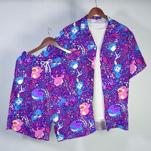May include: A matching purple short-sleeve button-down shirt and shorts set. The set features a vibrant pattern of cartoon characters in shades of purple, blue, pink, and white. The shorts have a drawstring waist. A white t-shirt is visible underneath the shirt.