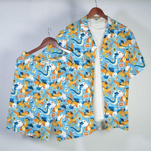 May include: A matching set of clothing, including a short-sleeved button-down shirt and shorts, both featuring a repeating pattern of orange and white cartoon dragons on a light blue background. A white t-shirt is visible underneath the shirt.