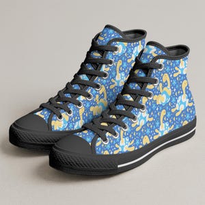 Könnte beinhalten: Hohe Canvas-Sneaker mit blauem Hintergrund und einem Muster aus gelben und blauen Zeichentrickschildkröten und Blumen. Die Sneaker haben schwarze Schnürsenkel, schwarze Sohlen und einen weißen Streifen entlang der Sohle.