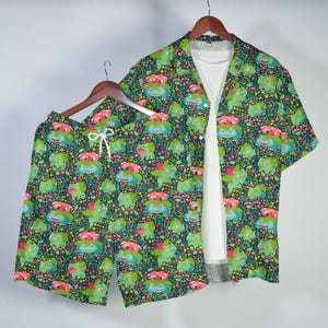 Puede incluir: Un conjunto de camisa de manga corta con botones y pantalones cortos a juego, con un fondo verde oscuro y un patrón repetitivo de criaturas verdes con flores rosas en la espalda, intercaladas con pequeños detalles florales amarillos y rosas.