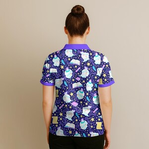 May include: A dark blue polo shirt with a repeating pattern of whimsical figures, books, stars, and feathers. The shirt has purple trim on the collar and sleeves. The overall design has a magical or fantasy theme.