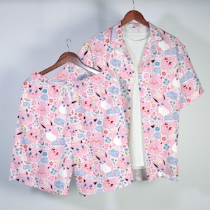 May include: A pink pajama set with a repeating pattern of cartoon characters, clouds, and flowers. The set includes a short-sleeved button-down shirt and matching shorts. A white t-shirt is visible underneath the shirt.