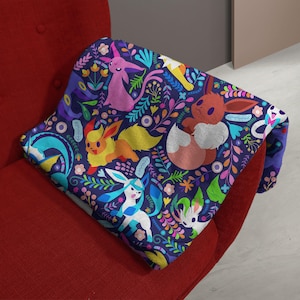 May include: A dark blue throw blanket featuring a repeating pattern of various colorful Eevee evolution characters, including Espeon, Flareon, Glaceon, and Leafeon, interspersed with vibrant floral and botanical elements in shades of pink, yellow, and green.