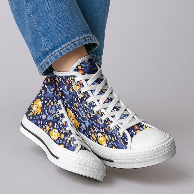 May include: High-top canvas sneakers with a navy blue background and a repeating pattern of yellow and blue cartoon dogs, flowers, and bones. The sneakers have white laces, a white toe cap, and a black stripe along the sole.