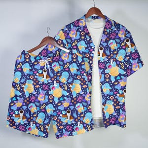 Puede incluir: Un conjunto a juego de pantalones cortos azules y una camisa de manga corta. Ambos artículos presentan un patrón repetido de personajes de dibujos animados, corales y elementos acuáticos sobre un fondo azul oscuro. La camisa está abierta, revelando una camiseta interior blanca.