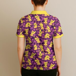 May include: Purple polo shirt featuring a repeating pattern of yellow cartoon characters. The shirt has a yellow collar and sleeve trim. The design includes yellow cartoon characters, flowers, and clouds on a purple background.