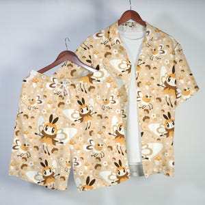 Può includere: Un completo pigiama beige con camicia a maniche corte e pantaloncini, decorato con un motivo ripetuto di falene, funghi e stelle dei cartoni animati. La camicia è aperta, rivelando una maglietta bianca.