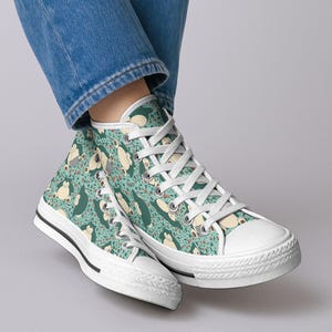 Op de afbeelding: Hoge canvas sneakers met een herhaald patroon van een stripfiguur in groen-, beige- en roodtinten op een turquoise achtergrond. De sneakers hebben witte veters, een witte neus en een zwarte streep langs de zool.