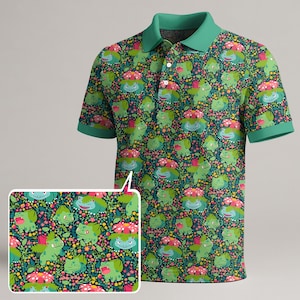 Op de afbeelding: Een poloshirt met korte mouwen, een effen groene kraag en mouwboorden. De stof heeft een all-over herhalend patroon van groene wezens met roze bloemen op hun rug, afgewisseld met kleine gele en roze bloemaccenten.