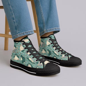 Op de afbeelding: Hoge sneakers met een teal achtergrond en een herhalend patroon van een stripfiguur in wit, beige en donkergroen. De sneakers hebben zwarte veters, een zwarte neus en een zwarte zool met een witte streep.
