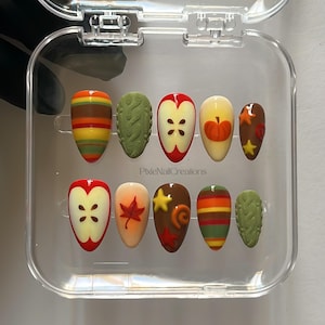 May include: A set of ten artificial nails in a clear plastic case. The nails feature autumn-themed designs, including apples, pumpkins, leaves, and stripes, in shades of brown, orange, yellow, and green. The brand name "PixieNailCreations" is visible.