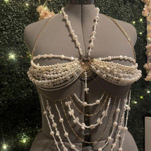 以下が含まれることがあります： マネキンに展示された、手作りの真珠とゴールドチェーンのボディハーネス。ハーネスは、白い真珠、ゴールドチェーン、装飾的なリボンの複数のストランドが特徴です。ハート型のペンダントが中央から吊り下げられています。